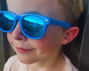 May include: Bright blue sunglasses with reflective lenses. The sunglasses are worn by a person. The lenses reflect a landscape with a road and buildings. The frames are a solid, vibrant blue.
