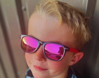 May include: A child wearing sunglasses with a black frame and red arms. The lenses are a vibrant pink, reflecting a sunset scene. The child has blonde hair and is wearing a striped shirt.
