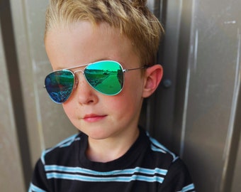 May include: A young person wearing silver-framed aviator sunglasses with green mirrored lenses. The lenses reflect a vibrant outdoor scene. The person is wearing a black and blue striped shirt.