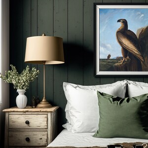 May include: A bedroom interior with a dark green wood wall, a white bed with white and green pillows, a bedside table with a lamp and a vase of flowers, and a framed painting of a brown eagle on a rock.