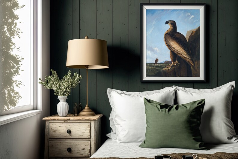 May include: A bedroom interior with a dark green wood wall, a white bed with white and green pillows, a bedside table with a lamp and a vase of flowers, and a framed painting of a brown eagle on a rock.