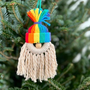 Op de afbeelding: Een handgemaakte kerstversiering in de vorm van een kabouter. Het heeft een regenboogkleurige hoed en een crèmekleurige baard van macramé. Een houten kraal dient als neus. De versiering hangt aan een kerstboom.