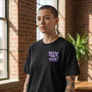 Könnte beinhalten: Schwarzes T-Shirt mit dem lila Schriftzug "BREAK THE BINARY". Der Text ist in einer stilisierten Schriftart gehalten. Das T-Shirt wird von einer Person mit kurzen Haaren getragen.