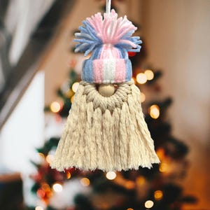 May include: A handmade gnome ornament with a cream-colored beard and a blue, pink, and white knit hat. The gnome has a wooden bead nose and a fluffy pom-pom on top of the hat. The ornament is hanging from a white string.