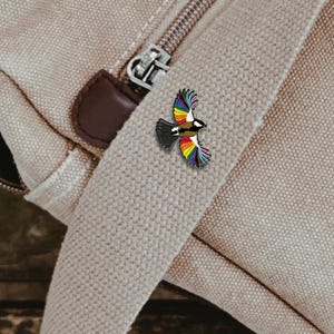 May include: A colorful bird pin with rainbow wings, black and white body, and a brown chest. The pin is attached to a beige canvas bag with a zipper and brown leather accents. The bird is in mid-flight.
