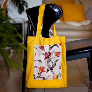 Può includere: Una borsa tote gialla con una stampa floreale con tulipani rossi, gialli e bianchi. La borsa è appesa a una sedia nera.