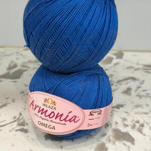 May include: A ball of vibrant blue mercerized cotton yarn with a light pink paper label wrapped around the center. The label features the text Armonía, Hilaza, Omega, 100% Algodón Mercerizado, and additional product information in black text.