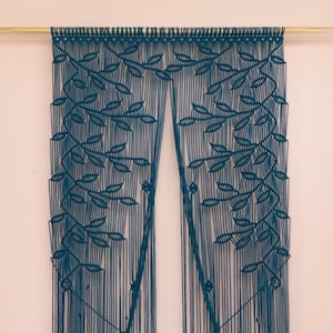 Puede incluir: Tapiz de pared de macramé color turquesa con diseño de hojas. La obra de arte textil está suspendida de una barra de madera y cuelga en dos paneles. El tapiz de pared se exhibe en una pared rosa. El texto "Dewy Love" es visible.