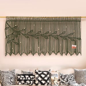 May include: A living room scene featuring a cream-colored sofa adorned with patterned pillows and a woven throw. A large, dark green macrame wall hanging with a leaf design hangs above the sofa. A wooden coffee table and patterned rug complete the decor.