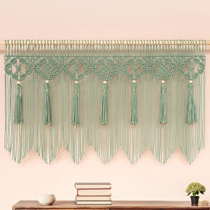 May include: A sage green macrame wall hanging with a scalloped bottom edge and tassels. The wall decor is suspended from a wooden dowel. Below, a wooden cabinet with a round jute rug and a potted plant complete the bohemian-style room.