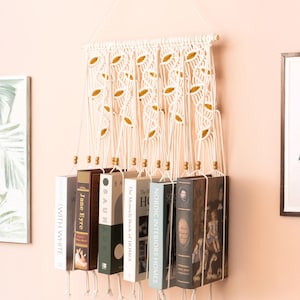 May include: A cream-colored macrame wall hanging with a leaf design, holding several books. The books are various colors and sizes, suspended by knotted cords. The wall is painted a light pink, with framed artwork visible.