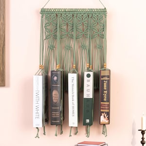 May include: A sage green macrame wall hanging with five books suspended from it. The books have various covers and titles, including "With White" and "Jane Eyre." The wall hanging is mounted on a light pink wall.