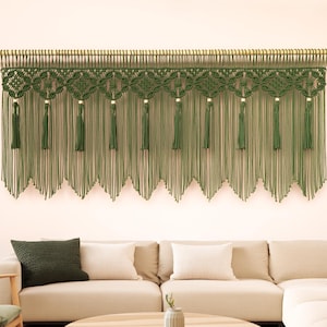 May include: A green macrame wall hanging with a wooden dowel. The macrame features intricate knotting patterns and long, flowing tassels. The wall decor is displayed above a beige sectional sofa in a modern living room setting.
