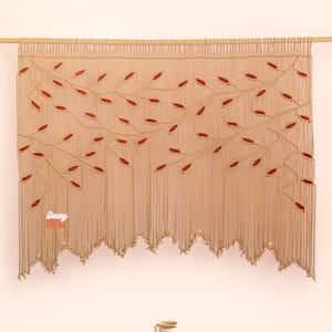 Op de afbeelding: Een beige macramé wandkleed met een rood bladeren ontwerp. De wanddecoratie is opgehangen aan een houten roede. De onderste rand heeft franjes. Het woord "Dewy" is linksonder zichtbaar. De kamer heeft een vloerkleed, een salontafel en planten.