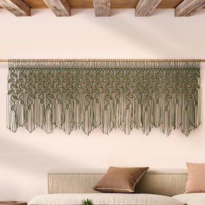 Może przedstawiać: Duża makrama ścienna w kolorze oliwkowym i beżowym, zawieszona na drewnianym drążku. Wzór przedstawia powtarzający się motyw liści i długie frędzle, dodając pomieszczeniu stylu boho. Makrama jest eksponowana nad beżową sofą.