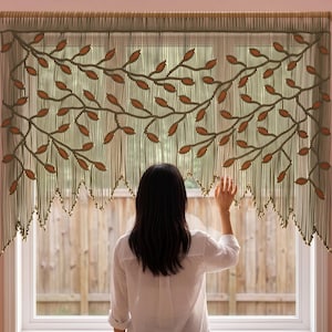 May include: A macrame window valance featuring a repeating vine pattern in olive green cord with integrated burnt orange leaf accents. The design includes a series of vertical hanging strands that terminate in a pointed, uneven fringe edge.