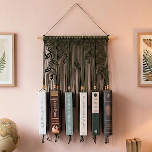 Puede incluir: Un tapiz de macramé verde oscuro con una varilla de madera y un colgador triangular. Seis libros cuelgan del macramé, incluyendo títulos como Jane Eyre, Nordic Interiors Homes y The Monocle Book of Homes.