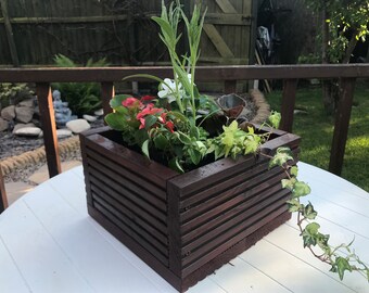 May include: A dark brown wooden planter box with a variety of plants, including green ivy, white flowers, and red flowers. The planter is sitting on a white table.