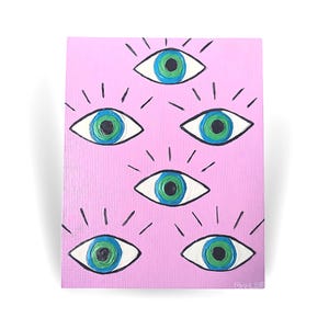 May include: A pink wooden panel featuring a repeating eye design. Each eye is white with a blue and green iris and black radiating lines. The artwork has a handmade, folk art style.