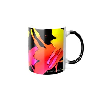 Puede incluir: Taza de cerámica blanca con un diseño abstracto colorido. El diseño presenta salpicaduras de amarillo, naranja y rosa sobre un fondo negro. La taza tiene un asa negra y el texto "Desert Studio Art Co."