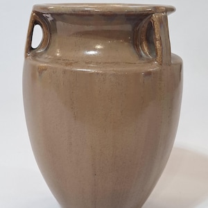 May include: A brown ceramic vase with two loop handles. The vase has a wide body that tapers to a narrow base. The vase is glazed with a matte finish and has a slightly textured surface. The vase is a neutral color that would complement any decor.