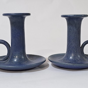 May include: Two blue ceramic candle holders. Each has a tall, cylindrical body with a flared top and a circular handle. The base is a wide, shallow dish. The candle holders are a matte finish.