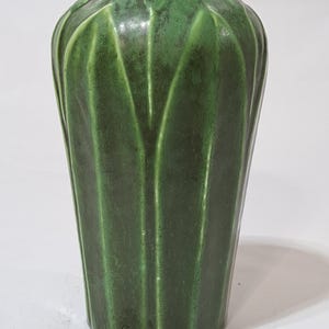 May include: A tall, green ceramic vase with a textured, ribbed design. The vase has a slightly flared top and a cylindrical body. The green color is a deep, rich shade, and the surface has a glossy finish. The vase is likely used for decorative purposes.