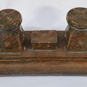 May include: Antique copper inkwell with two ink pots and a central compartment. The ink pots have domed lids with a cross design. The inkwell has a hammered texture and a rectangular base with a small trough.