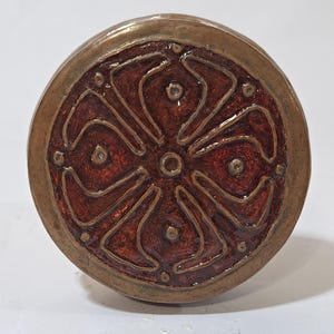 May include: A round, bronze-colored knob with a decorative, reddish-brown enamel design. The design features a central circle with a cross-like pattern, and small, circular accents. The knob has a metallic sheen and is likely for furniture or cabinetry.