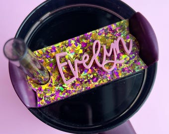 May include: A black tumbler lid featuring a rectangular resin name tag with the name Evelyn in raised light pink script lettering. The tag is filled with iridescent gold and purple glitter flakes and includes a clear straw insert.