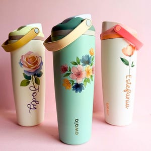 Puede incluir: Tres botellas de agua aisladas con diseños florales y asas. Una botella es de color crema con una rosa, otra es verde menta con un ramo, y la tercera es de color crema con una sola flor. Cada botella tiene un nombre.