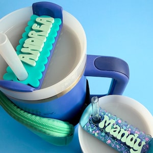 A blue and white sippy cup with a personalized name tag attached to it, sitting on a blue surface.