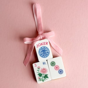May include: Three white rectangular mahjong tiles tied together with a light pink velvet ribbon. The top tile features the word JOKER in red with a blue circular design. The bottom tiles show a pink chrysanthemum flower and three colored circles.