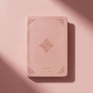 May include: A light pink, rectangular journal with gold embossed floral corner and center designs. The journal has the name "Lilian M." and the date "01.02.1990" embossed in gold on the bottom.