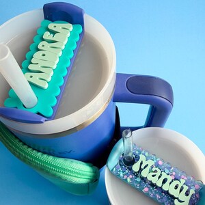 A blue and white sippy cup with a personalized name tag attached to it, sitting on a blue surface.