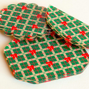 May include: Three acrylic coasters featuring a green and beige plaid pattern with small red bows printed throughout. The set includes one scalloped circular coaster, one semi-circular coaster, and one octagonal coaster with rounded edges.