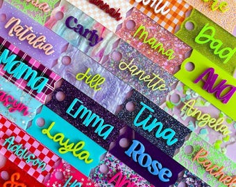 May include: A collection of rectangular acrylic tags with rounded corners and a circular hole, each featuring a raised 3D name. The tags display various patterns including glitter, checkered, plaid, and marble designs in diverse colors.