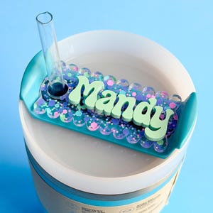 A colorful, personalized nameplate with the name "Manay" displayed on it, placed on top of a white container or cup.