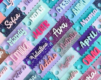 May include: A collection of rectangular acrylic tags with scalloped edges, each featuring a raised 3D name in a cursive or print font. The tags come in various glittery and solid colours including purple, blue, pink, and silver.