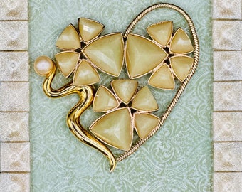 May include: A gold heart-shaped brooch with a pearl accent. The brooch is made of metal and has a design of interlocking triangles. The brooch is set against a green background.