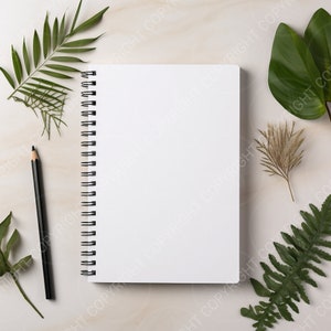 May include: A white spiral bound notebook with blank pages, a black pencil, and green leaves on a white marble surface.