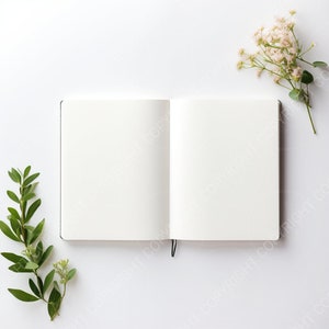 Puede incluir: Un cuaderno blanco abierto con páginas en blanco sobre un fondo blanco con ramitas verdes y flores blancas a cada lado.