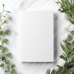 May include: A white blank book cover mockup on a marble background with white flowers and green leaves on either side.