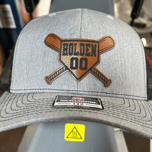 May include: A gray and black baseball cap with a brown leather patch featuring the name "Holden" and the number "00" inside a baseball diamond with two crossed baseball bats.