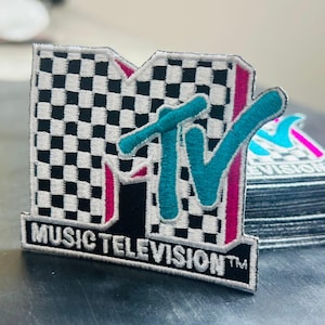 May include: Embroidered patch featuring the MTV logo. The patch has a black and white checkered background, teal "MTV" lettering, and pink accents. The words "MUSIC TELEVISION" are embroidered below the logo. A stack of similar patches is visible.