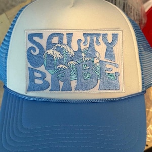 May include: A white and blue trucker hat with a blue embroidered patch that says "Salty Babe" with a wave design. The hat has a blue mesh back.