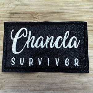 May include: Black embroidered patch with white text that reads "Chancla Survivor".