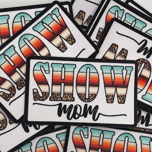 May include: Rectangular white patch with the words "SHOW MOM" in a colorful design. The word "SHOW" has a turquoise, orange, and brown striped pattern with leopard print accents. The word "mom" is in a black script font.