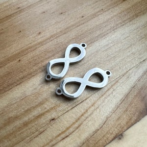 Könnte beinhalten: Zwei silberne Unendlichkeits-Anhänger mit einem Loch oben zum Anbringen an Schmuck oder anderen Bastelarbeiten.
