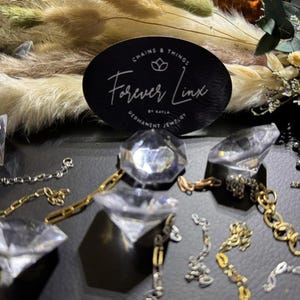 May include: A close-up of various silver and gold chains and charms on a black surface. The chains are arranged in a decorative pattern. A sticker with the text "Chains & Things Forever Linx by Kayla Permanent Jewelry" is visible in the background.